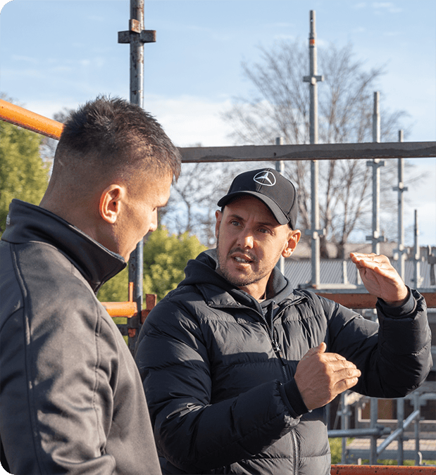 Two men in jackets engage in a discussion at a bustling construction site. One gestures animatedly, supercharging the dialogue, while the other listens attentively. Metal scaffolding and trees stand tall in the background, offering a glimpse into their thriving trade business.